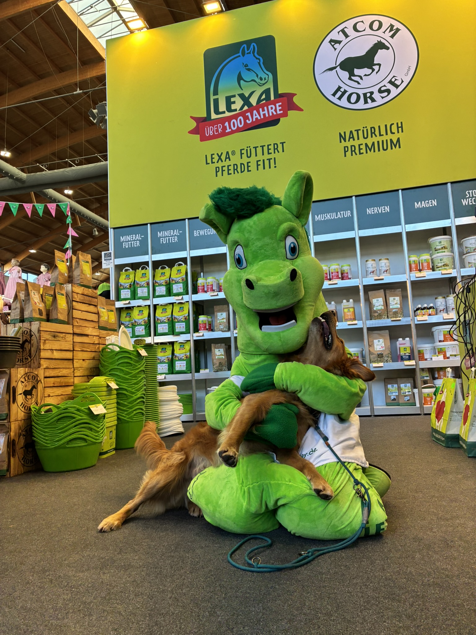 LEXA Maskottchen kuschelt mit Hund vor großem Messestand mit Pferdefutter-Produkten